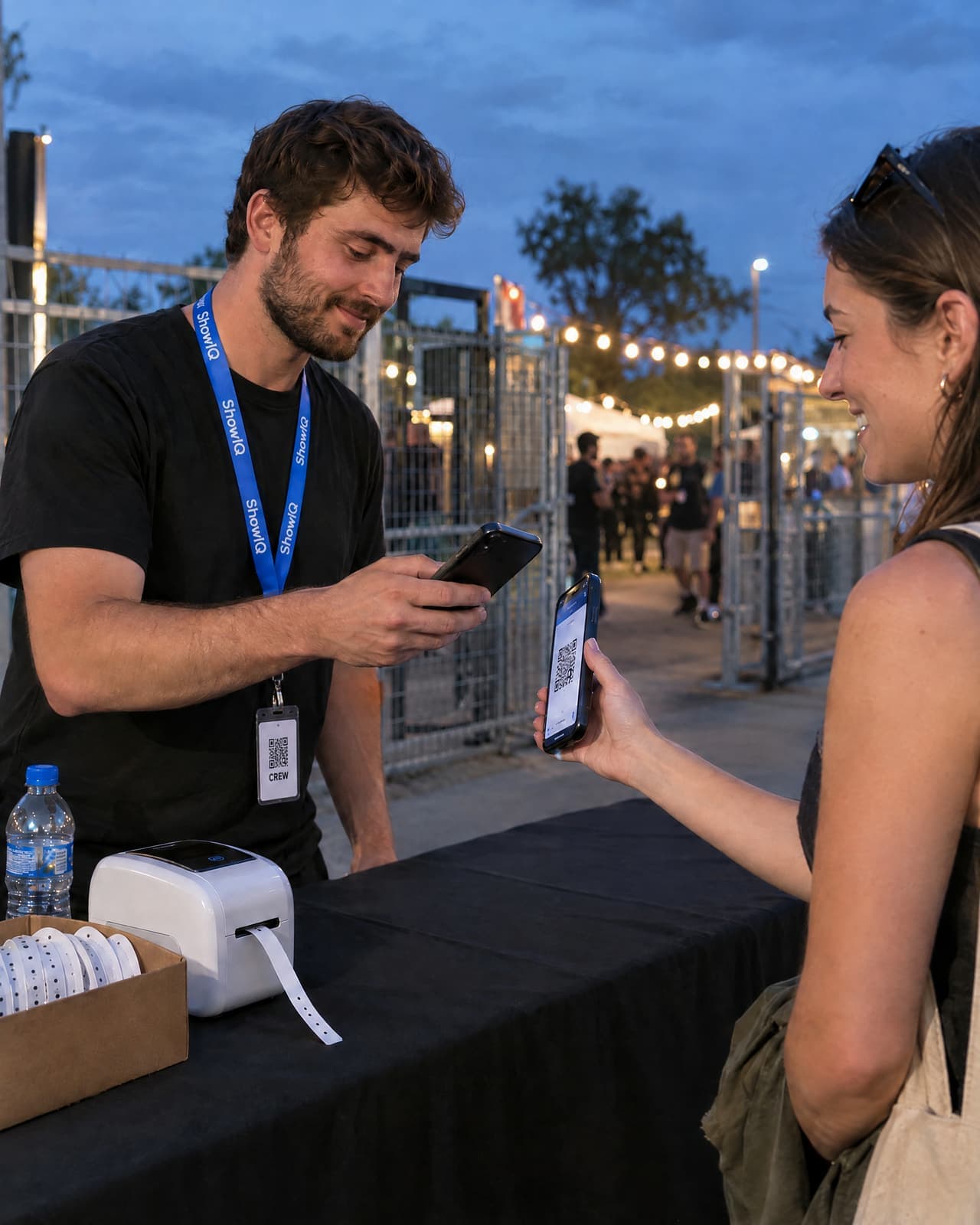 Door staff at the accreditation desk scanning the QR ticket on a visitor's phone, a wristband printer on the table
